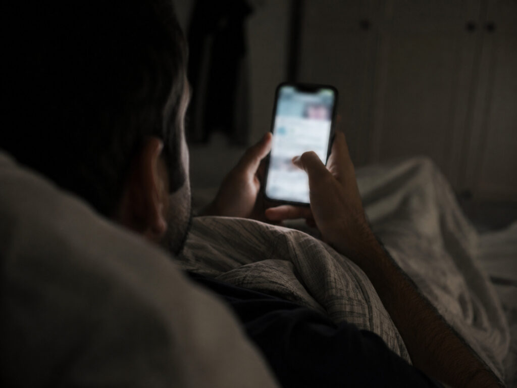 Person lying in bed at night looking at a glowing smartphone screen in a dark room.